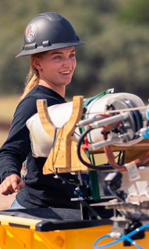 A person wearing a helmet and safety vest smiles while working with mechanical equipment mounted on a yellow structure outdoors.