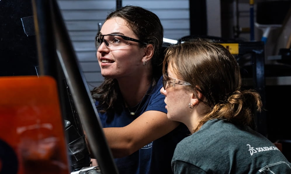 Two people wearing safety goggles work on mechanical equipment in a laboratory.