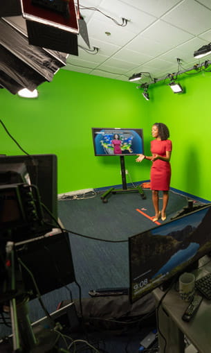 A television studio with a green screen where a presenter stands in front of weather monitors, surrounded by cameras, lights, and production equipment.
