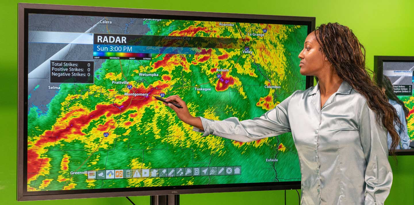 Meteorology student pointing at Doppler weather radar display showing a severe storm line over central Alabama, including Montgomery, Auburn, and Tuskegee, on a large touchscreen monitor in a studio setting.