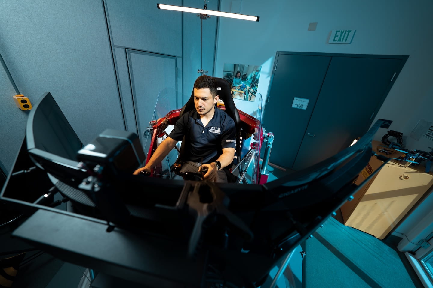 Student seated in a motion-based flight simulator rig, using dual control sticks and facing a multi-monitor display setup, with metal frame, actuators, and protective panels visible in a lab room.