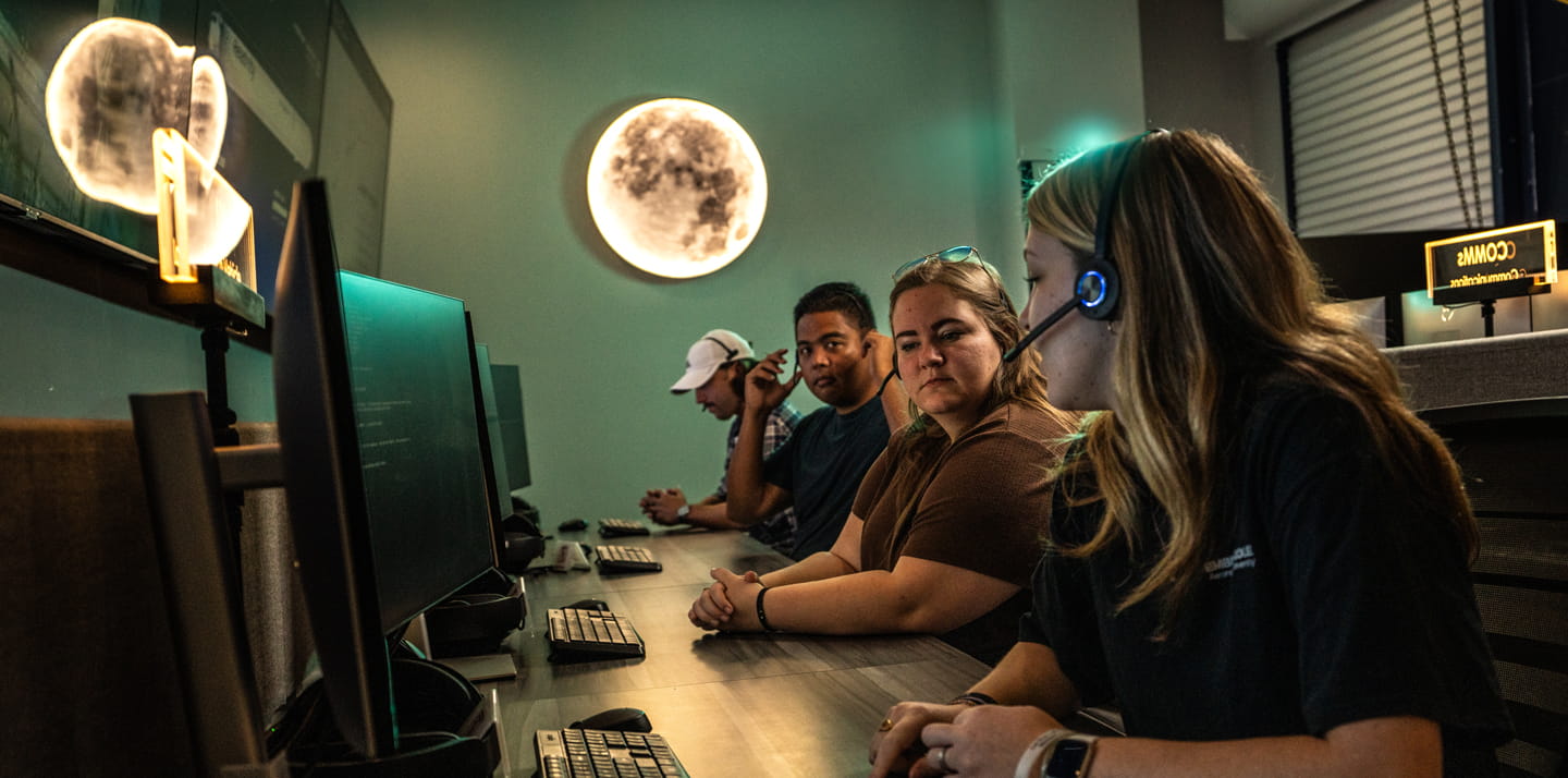 Students working at computer stations in Space Operations lab, one wearing headset and assisting others while code displays on monitors. A glowing moon wall decoration is hung on the wall behind them.