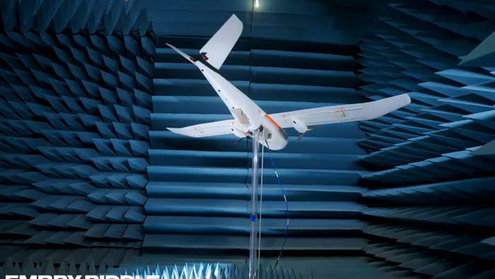 Fixed-wing unmanned aircraft mounted for testing inside anechoic chamber with acoustic foam panels on walls.