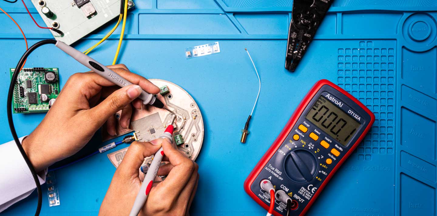 Hands use multimeter probes to test a printed circuit board on a workbench with electronic components and tools, including a digital multimeter and wire strippers.