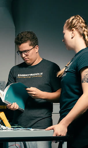 Two people stand at a workbench in a lab or workshop, reviewing documents and equipment together.