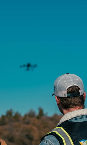 A person wearing a cap and high-visibility vest is seen from behind, looking up at a drone flying in the sky over a natural landscape.
