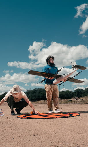 Two people on a sandy, open landscape prepare a small fixed-wing drone for flight.
