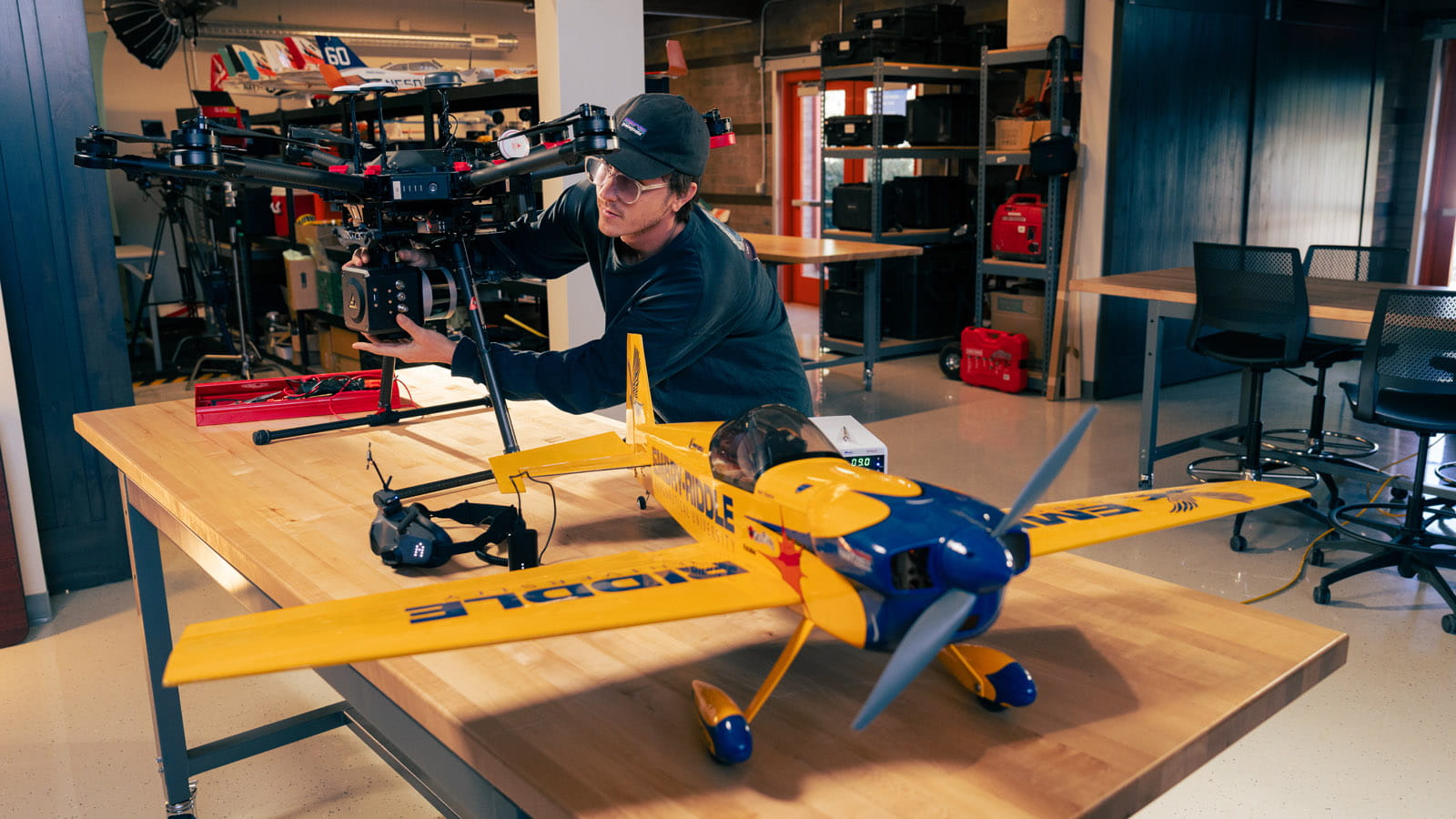 A person adjusts a large professional camera mounted on a multirotor drone inside a workshop, while a yellow-and-blue model airplane sits on a worktable in the foreground, with shelves of equipment and tools in the background.
