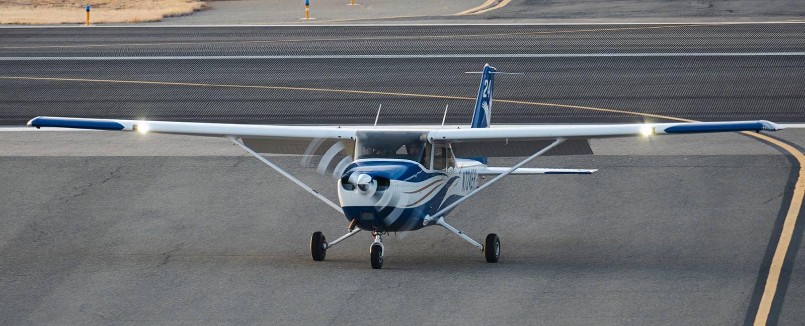 High-wing single-engine training aircraft with spinning propeller taxiing toward camera on runway marked 21, nose wheel steering visible and landing lights on, with taxiway lines and dry grass in background.