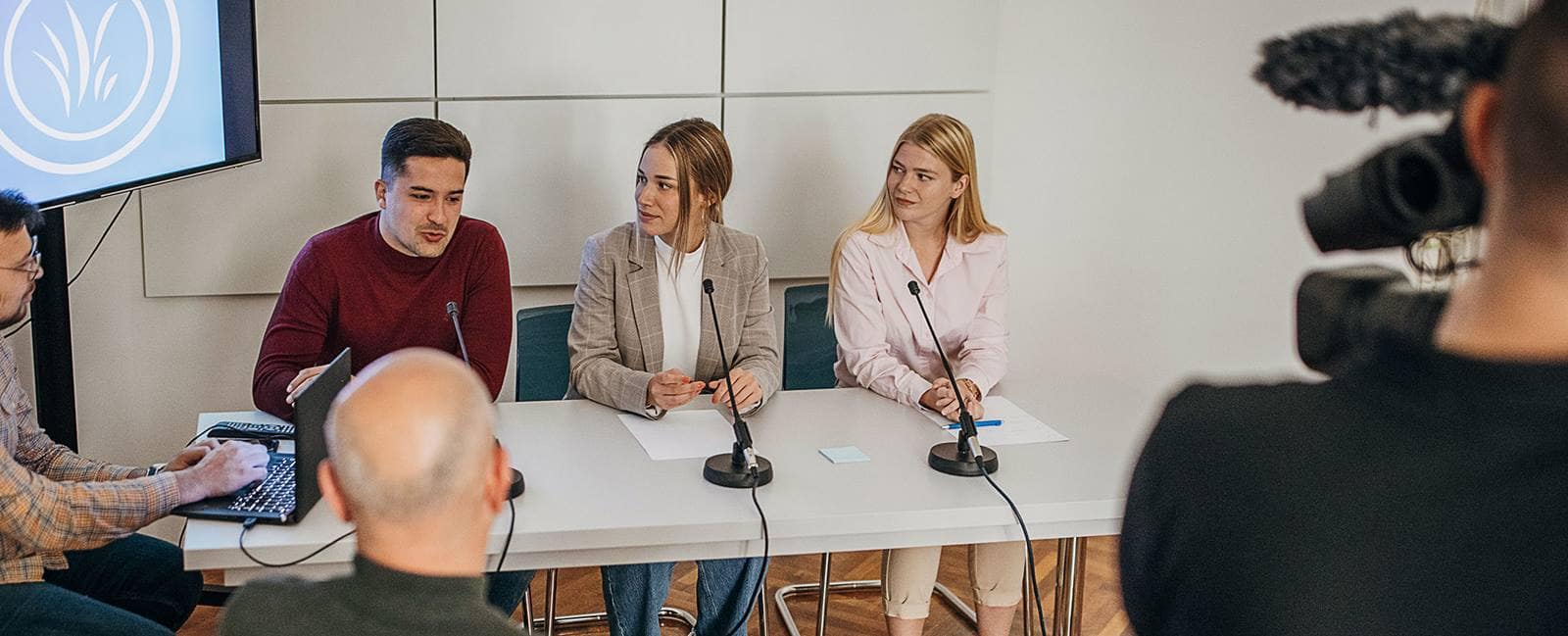 Three students seated at a panel table speaking into desktop microphones during a recorded discussion, camera and operator visible in foreground and presentation screen behind them.