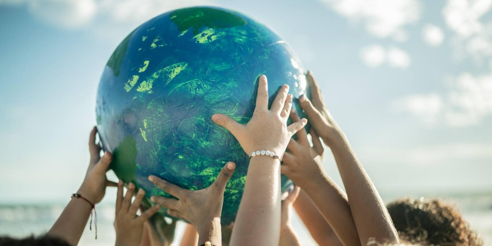 Multiple hands of diverse students lifting a beach ball with decoration like a globe, held up against bright sky near water.