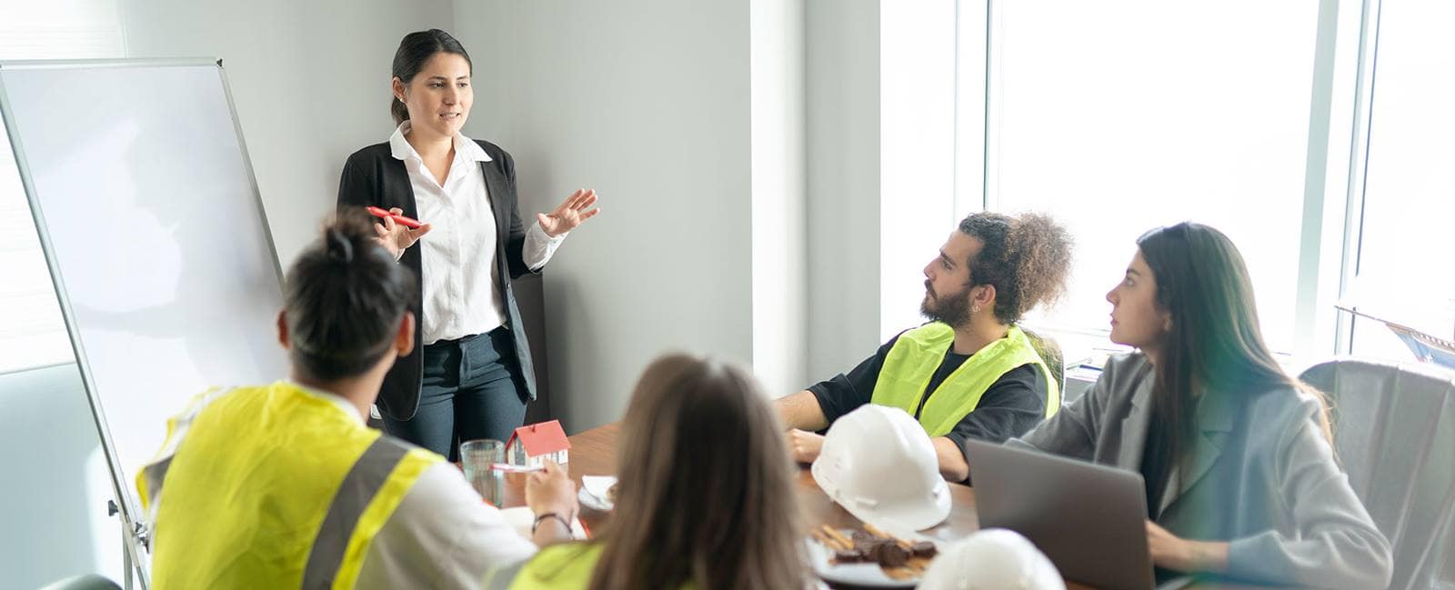 Presenter in business attire gestures while explaining to a small group of engineers and construction staff seated around a table with a laptop, hard hats, and a flip chart in a meeting room,.