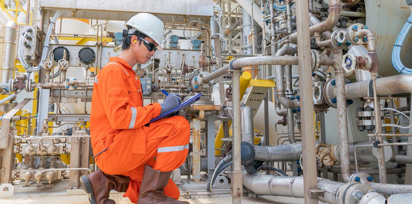 Technician in hard hat and protective gear kneels beside a dense network of industrial pipes, valves, gauges, and flanges, recording data on a clipboard during equipment inspection in a processing facility.