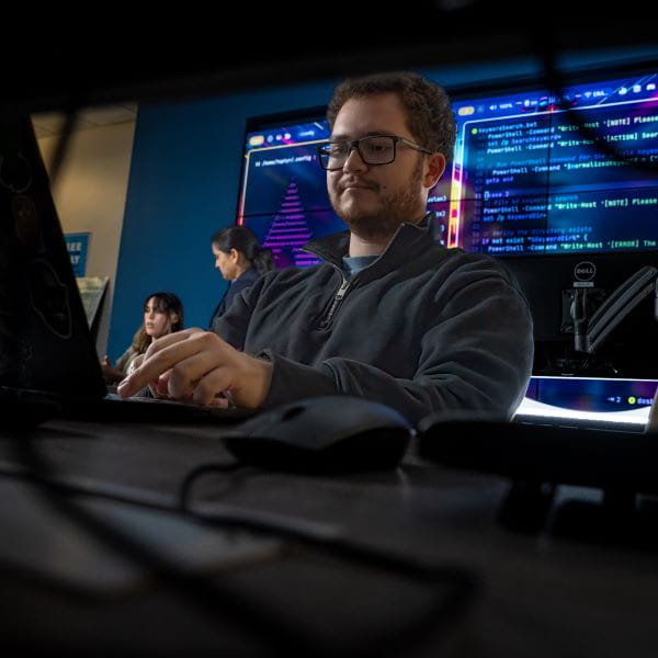 A person sits at a desk working on a computer in a control room setting.