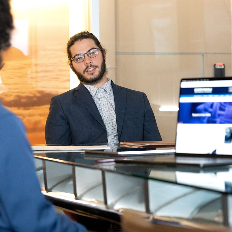 Two people sit across from each other in an office setting, having a conversation across a desk.