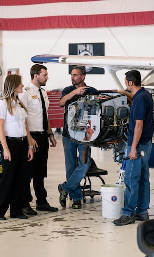 Students and instructor discussing aircraft engine mounted on stand in hangar with wing and maintenance equipment nearby.