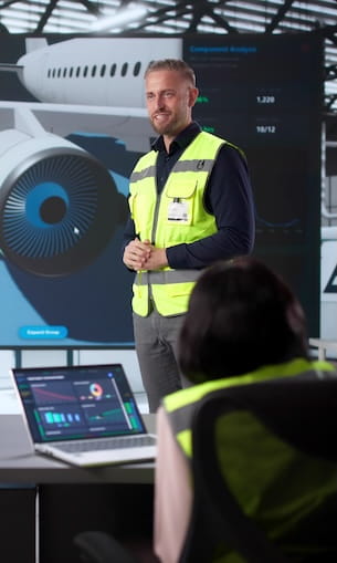 Instructor in safety vest presenting aircraft engine data and diagrams to students with laptop and display screen.