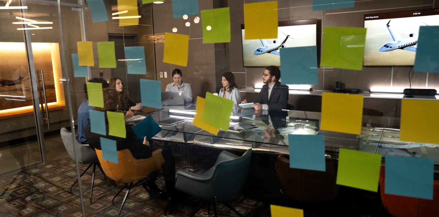 A group of people seated around a table in a glass-walled conference room, with colorful sticky notes posted on the walls.