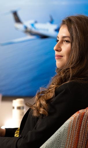 A person wearing business attire, sitting at a desk with a photo of an airplane in the background.