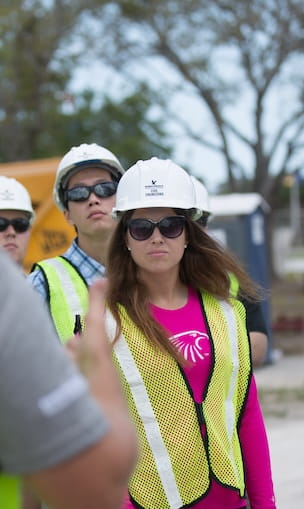 Students wearing hard hats and safety vests listening during construction site field training.