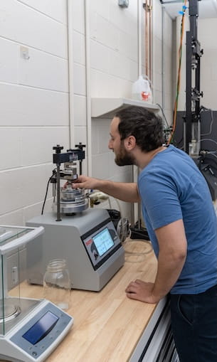 Student operating materials testing machine with cylindrical sample and digital controls in engineering lab.