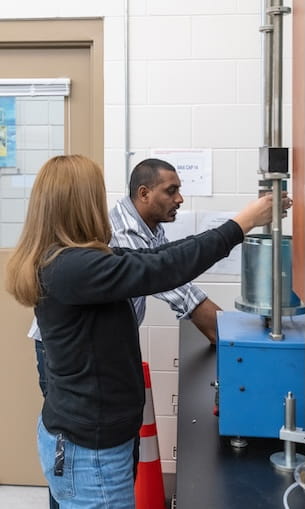 Students operating materials testing apparatus with vertical load frame and cylindrical sample in engineering lab.