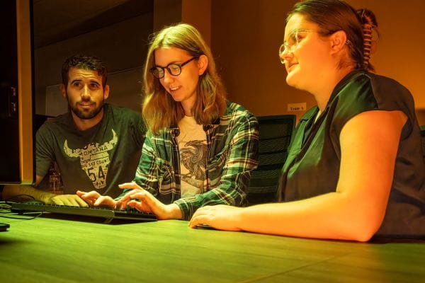 Three students sit at a desk working on a computer.