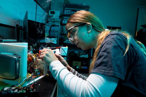 A student wearing glasses carefully works with electronic components on a circuit board in a lab setting.