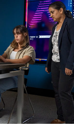 A professor watches as a student works at a computer.