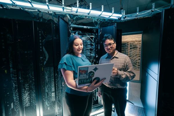 Two people stand together looking at a laptop with server equipment in the background.