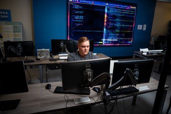 A person sits at a desk in a computer lab working on a computer with two monitors.