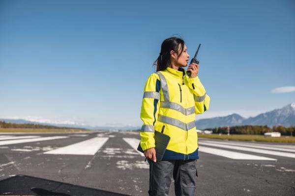 A person in a safety jacket stands on a runway, speaking into a radio.