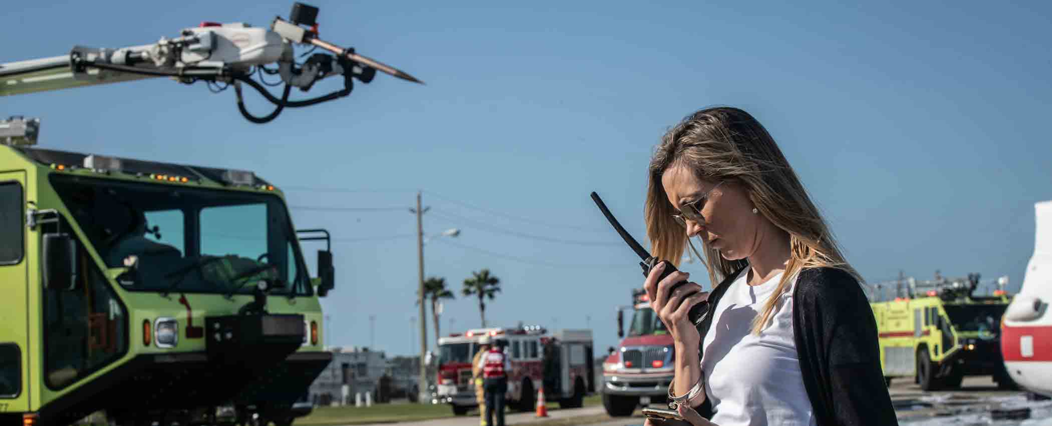 A person uses a walkie-talkie with a fire engine in the background.