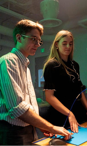 A male student and female student gaze intently at their work on a computer.