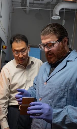 Two men, one in a blue lab coat, examine an electrical device.