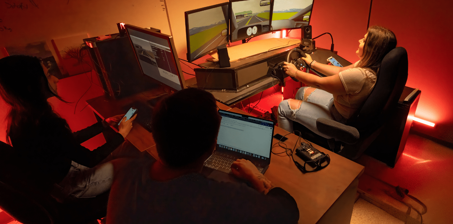 Students conduct a driving simulation study in a human factors lab, with one participant using a steering wheel and multi-screen roadway simulator while others monitor data on laptops and smartphones.