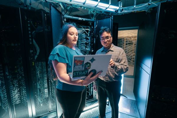 Two people standing side by side, looking at a laptop, with server equipment in the background.