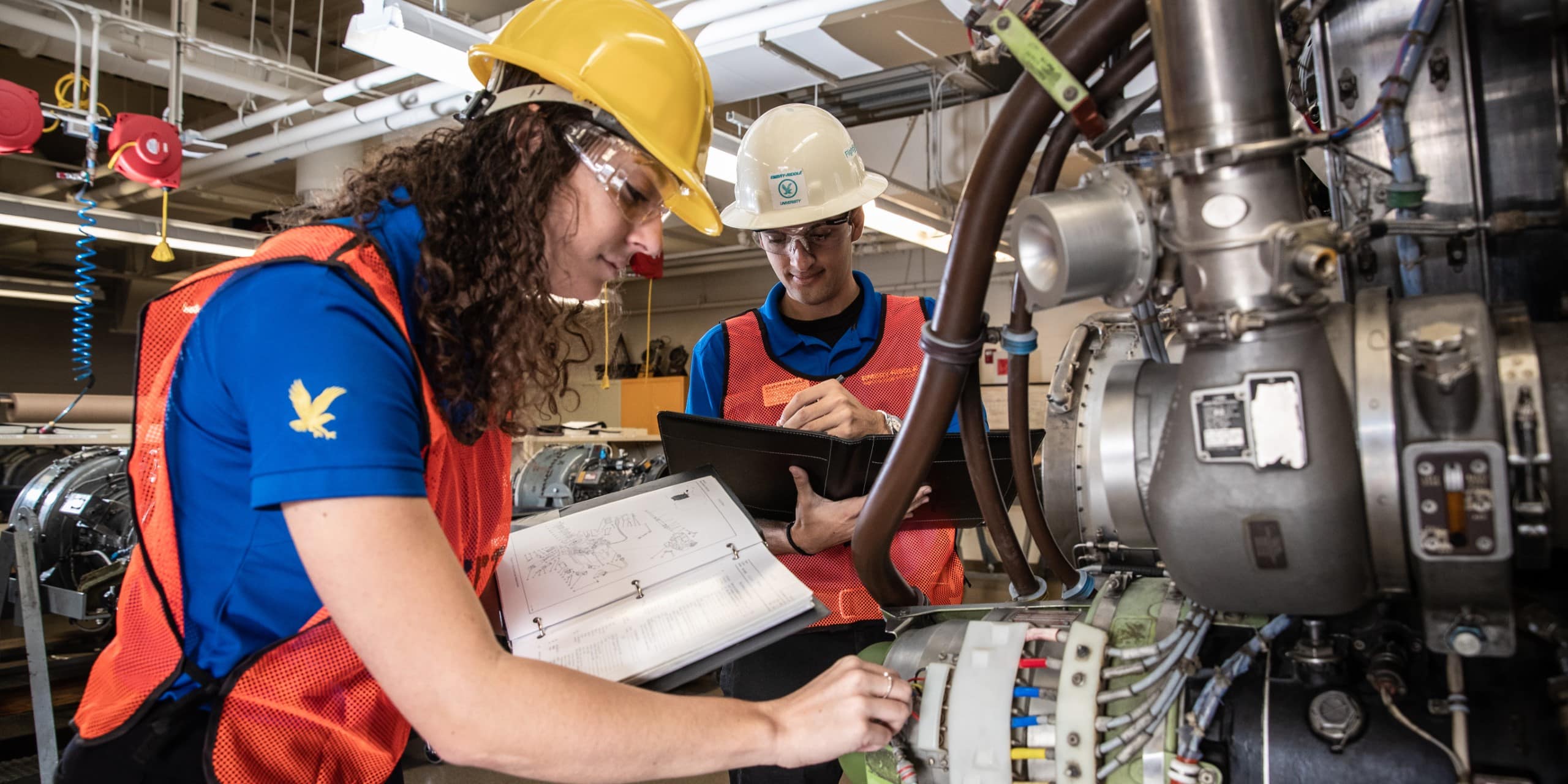 Two people wearing safety helmets and vests work on equipment in an industrial setting.