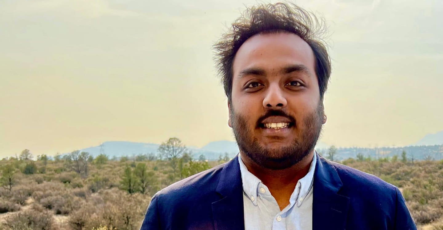 A person wearing a blue suit standing outdoors with mountains in the background.