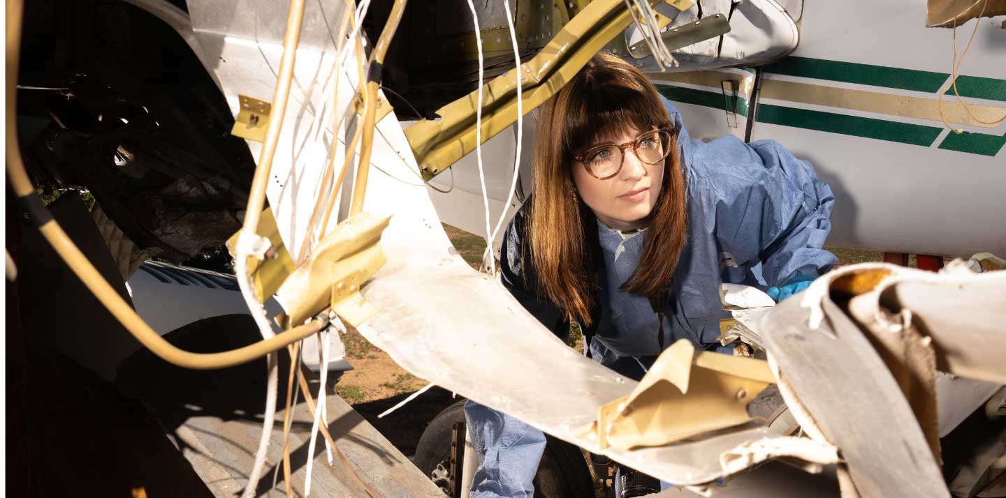 A woman wearing protective coveralls, gloves, and glasses is closely inspecting the damaged interior structure of a small aircraft.