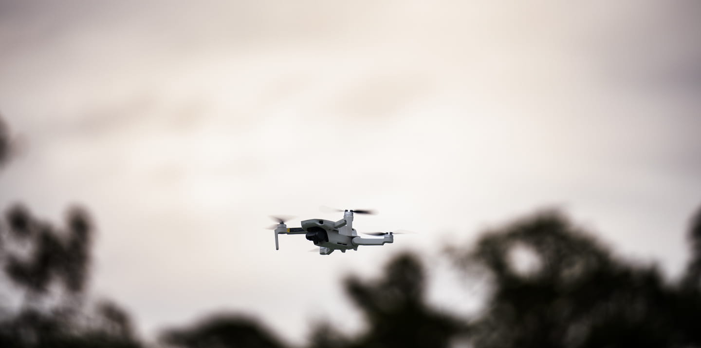 Drone hovering in front of a person wearing a safety vest and wide-brim hat in a wooded outdoor area.