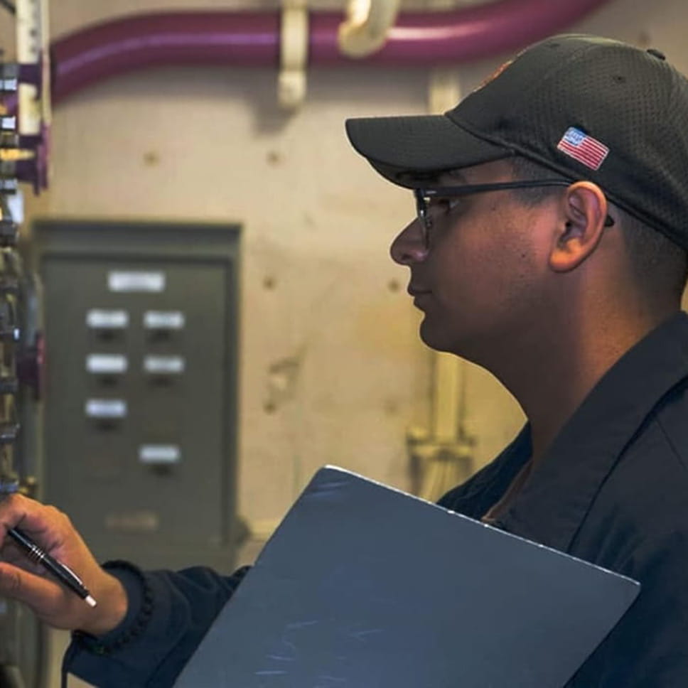 A person carrying a clipboard looks at a wall of switches and controls.