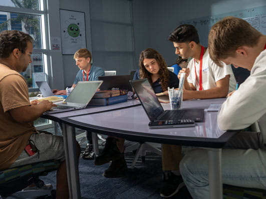 A group of people sitting around a table in a study or meeting space, working on laptops.