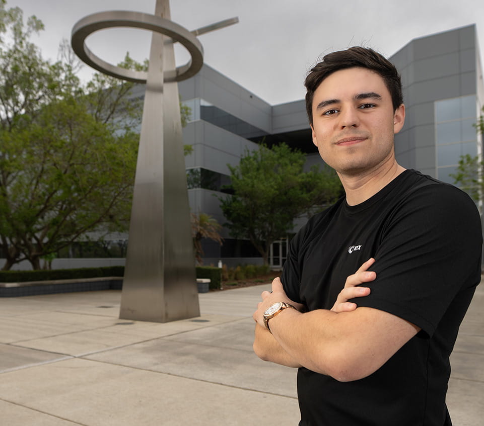 An individual stands outdoors in front of a building with their arms crossed.