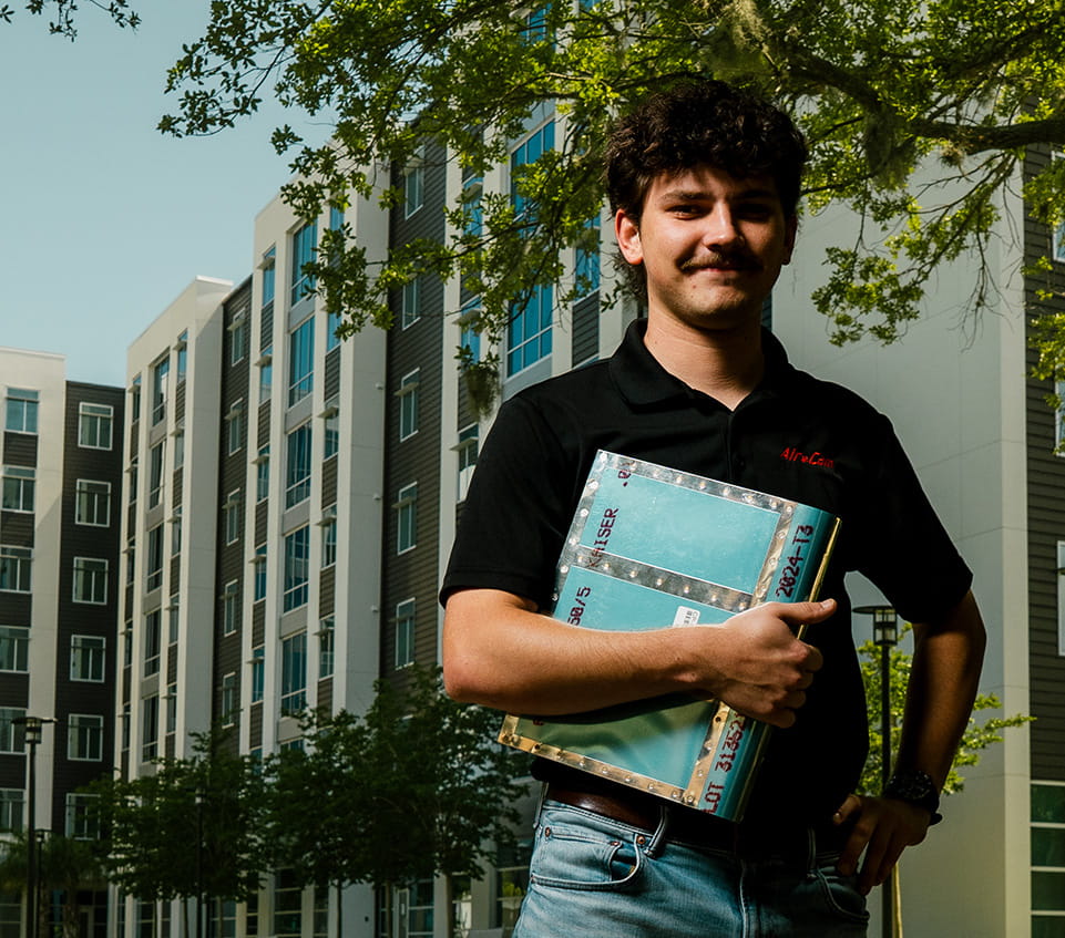 Student stands in front of residence halls, holding a binder.