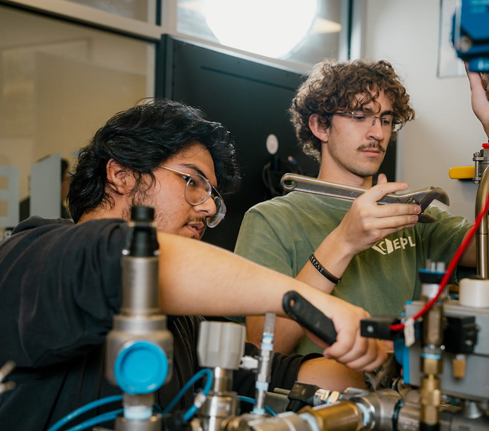 Two students adjust valves and fittings on a rocket propulsion test setup, one using a wrench while the other reaches across tubing and pressure regulators in a lab setting.