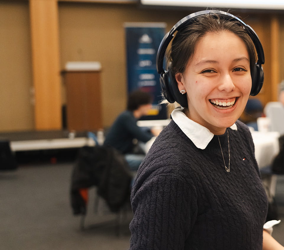 Student wearing headphones smiles broadly at the camera, other students at an event sit in the background.