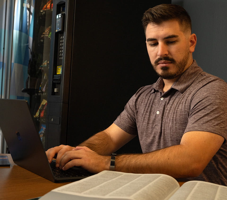 Student looks down at an open book while typing on a laptop.