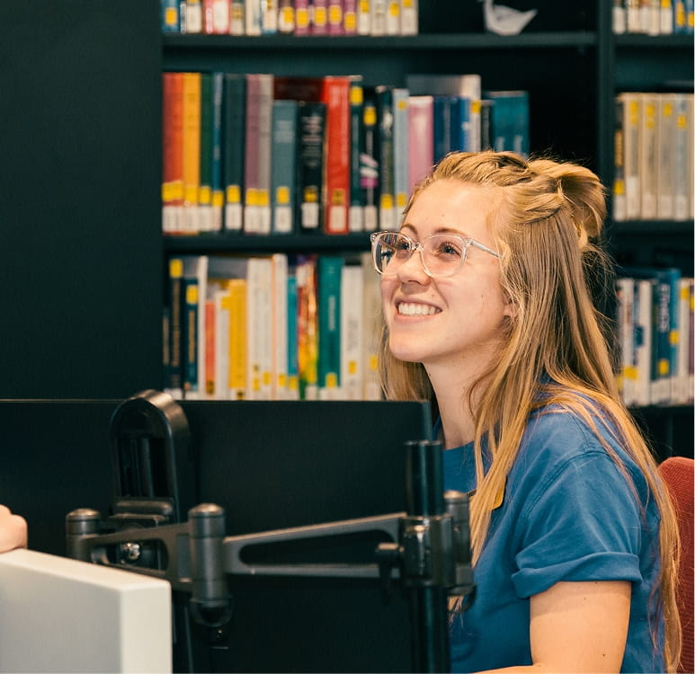 Student wearing glasses smiles while seated at a computer workstation, speaking with someone at a library help desk, with shelves of books in the background.