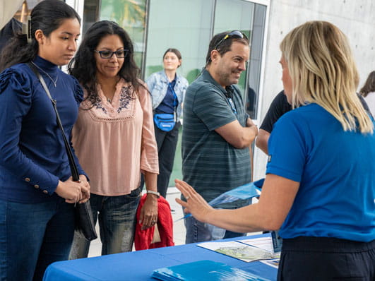 A group of people standing at an informational booth listening to a lady speak.